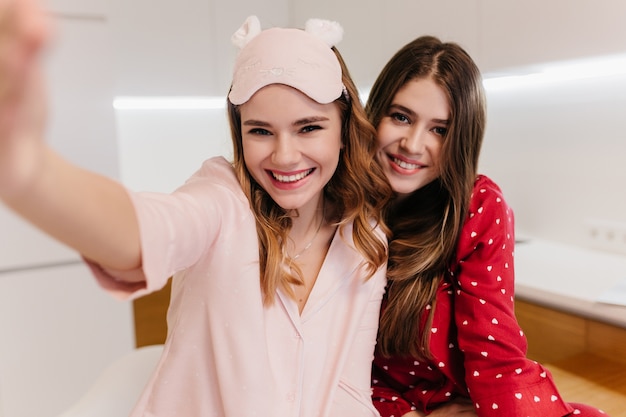 Dark-haired positive woman posing while sister making selfie. Dreamy young ladies smiling early in morning.