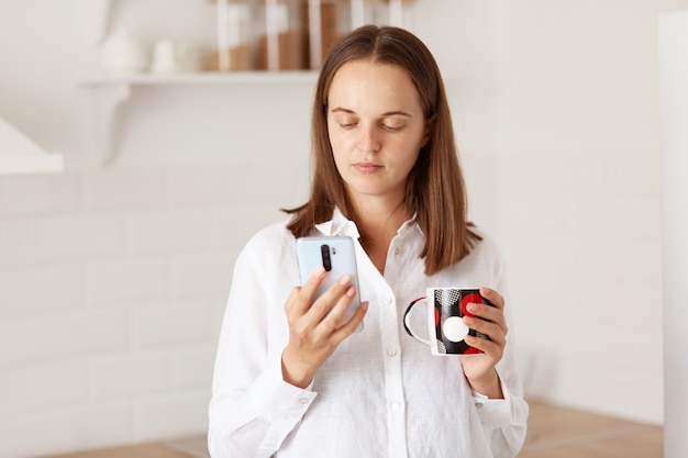 Dark haired woman standing with smart phone, reading news in social networks, enjoying hot coffee or tea in the kitchen in the morning, wearing white casual style shirt.