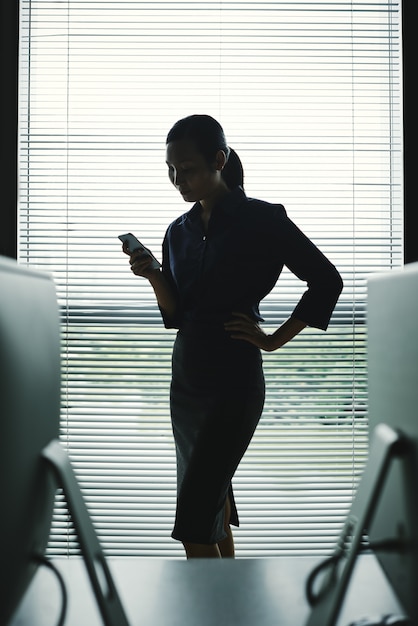 Dark silhouette of woman with smartphone standing in office at window with blinds