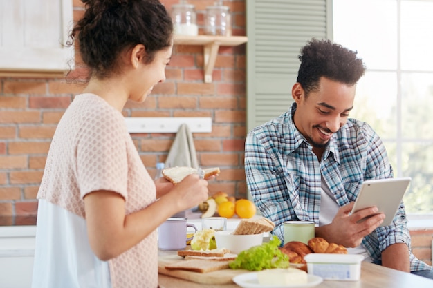 Free photo dark skinned afro american man dressed casually sits at kitchen with tablet computer, reads news online when his wife makes sandwhiches.