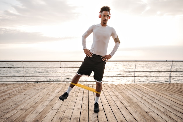 Free photo dark-skinned curly brunette man in white long-sleeved t-shirt and black shorts listens to music in headphones and does exercises with fitness rubber near sea