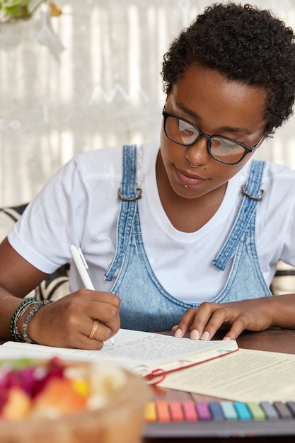 Dark skinned hipster in eyewear, writes in notebook, does homework 