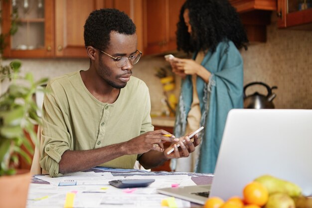 Dark-skinned man in spectacles doing finances, using cell phone, calculator and laptop computer