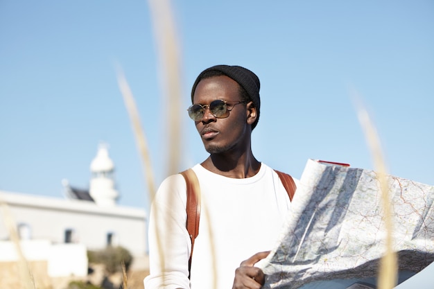 Dark-skinned traveler in trendy sunglasses and headwear studying paper map in his hands, looking worried while got lost during road trip, having concentrated expression, trying to find right direction