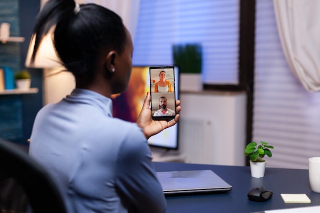 Dark skinned woman talking about project with coworkers late at night in the course of video conference on smartphone. Busy employee using modern technology network wireless doing overtime for job.