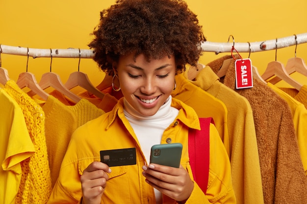 Free Photo dark skinned woman uses modern mobile phone and credit card, does online shopping, makes order via internet, inserts bank account information, stands against clothing racks.