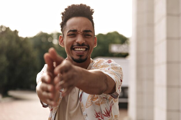 Darkskinned curly bearded charming man in white floral shirt talks looks into camera and poses outside