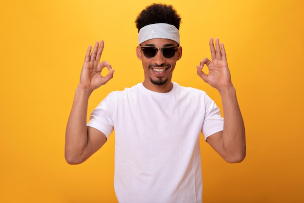 Free photo darkskinned guy in white tshirt and glasses shows sign ok on orange background cheerful man in tee and headband widely smiling on isolated