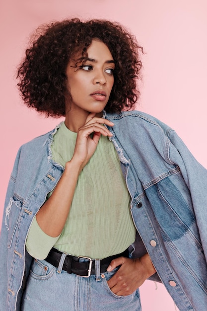 Darkskinned woman in jeans outfit and green Tshirt thoughtfully posing on pink background Portrait of lady in denim jacket and skirt on isolated backdrop