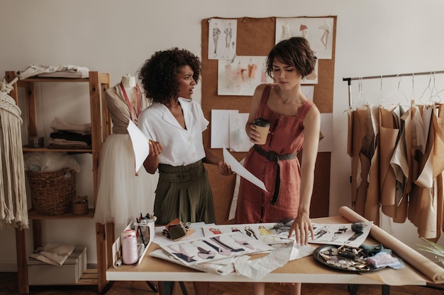 Darkskinner young curly woman in khaki shorts and white blouse holds paper sheets and talk to shorthaired girl in linen dress Women work as fashion designers and look dissatisfied