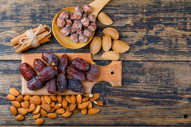 Dates on a cutting board with peeled and unpeeled almonds, nuts in wooden spoon, cinnamon sticks top view on a wooden background