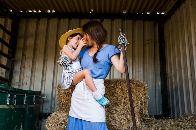 Free Photo daughter feeding red apple to her mother standing in front of hay stack