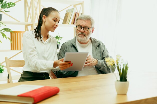 A daughter or granddaughter spends time with the grandfather or senior man. Family or fathers' day, emotions and happieness. Lifestyle portrait at home. Girl taking care about dad. Using a tablet.