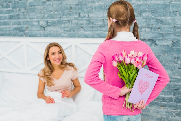 Free photo daughter holding greeting card and tulips for mother in bed