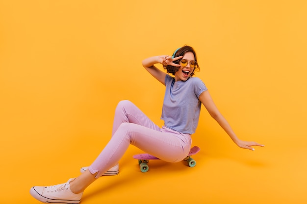 Debonair caucasian girl wears white shoes sitting on skateboard. Laughing pretty lady in headphones posing.