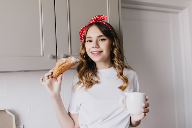 Debonair caucasian woman eating croissant with pleasure. Amazing glad girl drinking cappuccino in morning.