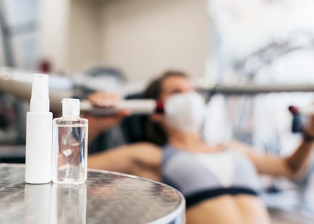 Defocused woman at the gym using equipment with medical mask and hand sanitizer bottle