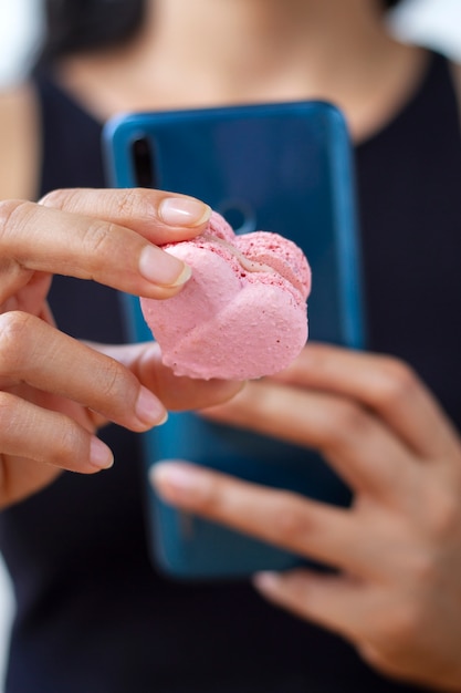 Defocused woman taking photo of heart-shaped macaron with smartphone