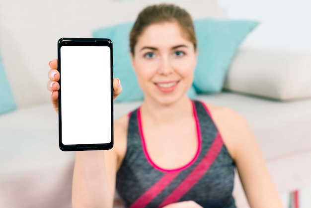 Defocussed young woman sitting near the sofa showing smartphone with blank white screen