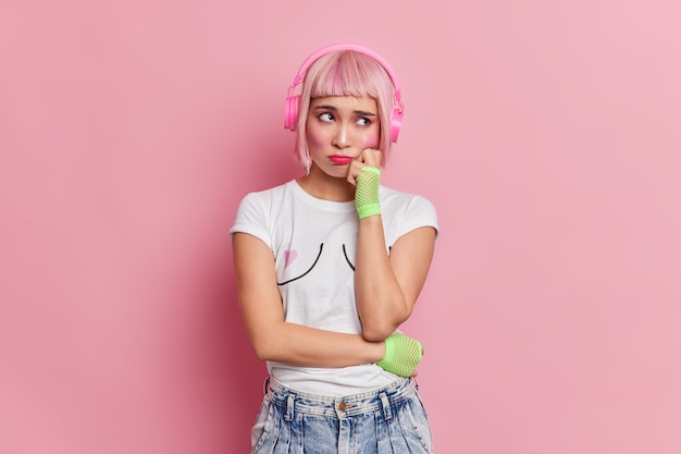 Free Photo dejected displeased asian woman has unhappy expression keeps hand on cheek wears casual t shirt jeans listens music via wireless headphones upset to have unlucky day 