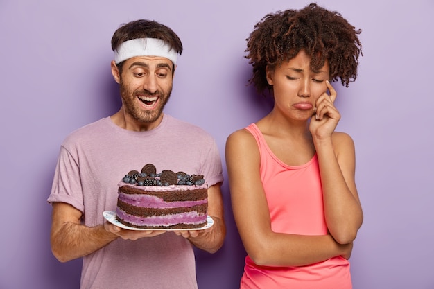 Free photo dejected upset woman turns from husband who holds tasty cake on plate, has sad expression as cannot eat sweet desserts for keeping fit and slim leads healthy lifestyle, refuses eating junk food