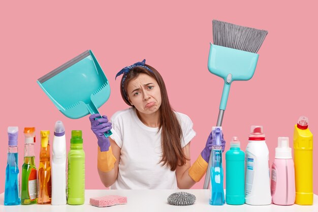 Dejected upset young housemaid frowns face in discontent, holds scoop and broom, does cleaning