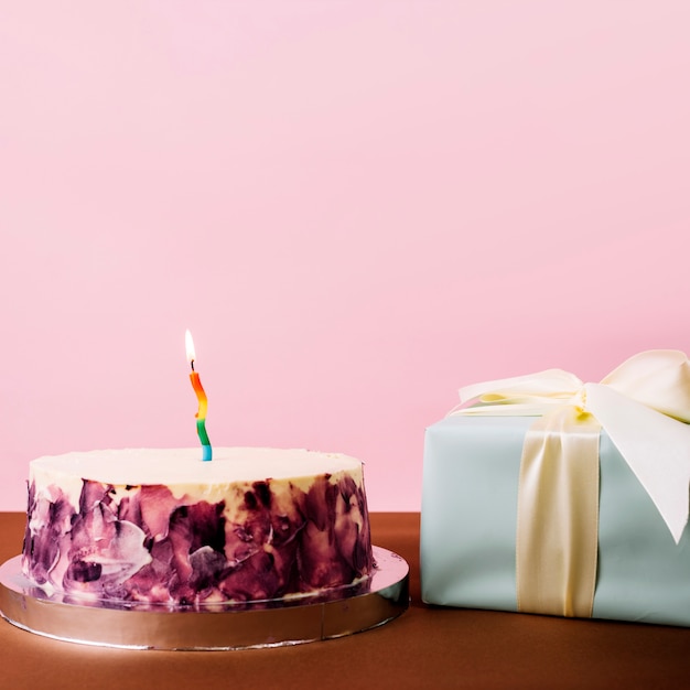 Free photo delicious cake with illuminated candle and wrapped gift box against pink background