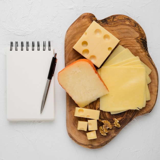 Delicious cheese platter and blank spiral dairy with pen against plain background