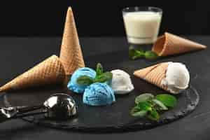 Free photo delicious creamy and blueberry ice cream decorated with mint, and waffle cones are served on a stone slate, standing on a dark table over a black background. metal scoop and a glass of milk is nearby.