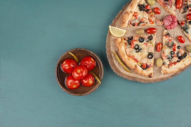 Delicious hot pizza and bowl of pickled tomatoes on blue table.