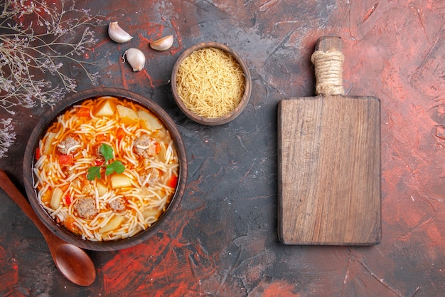 Delicious noodle soup with chicken and uncooked pasta in a small brown bowl and spoon garlic next to cutting board on the dark background