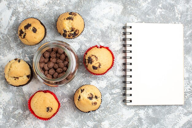 Above of delicious small cupcakes around chocolate cookies in a glass pot next to notebook on ice surface