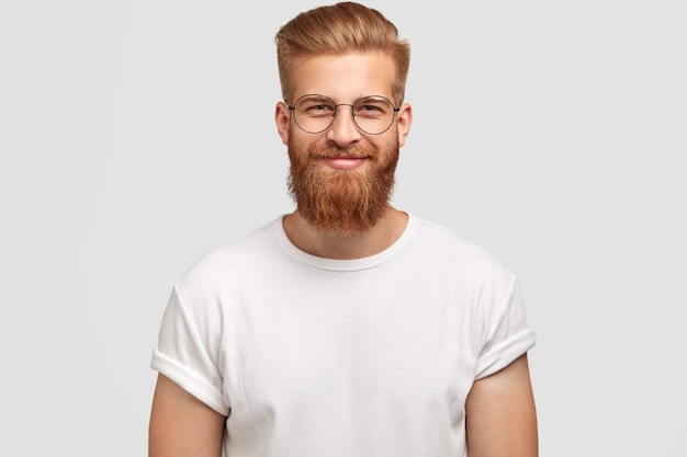 Delighted cheerful fashionable hispster with ginger beard, wears round spectacles and white t shirt