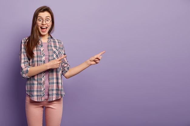 Delighted dark haired woman in checkered shirt, smiles broadly, indicates at copy space