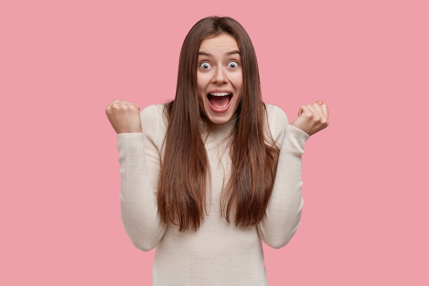 Free Photo delighted emotive european woman clenches fists, opens mouth and eyes, exclaims with victory mood, wears casual clothes, celebrates triumph