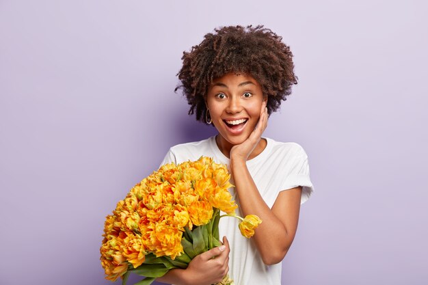 Delighted female with toothy smile, crisp hair, happy receive proposal from boyfriend, holds nice bouquet of yellow flowers, isolated against purple wall. Positive emotions and feelings concept