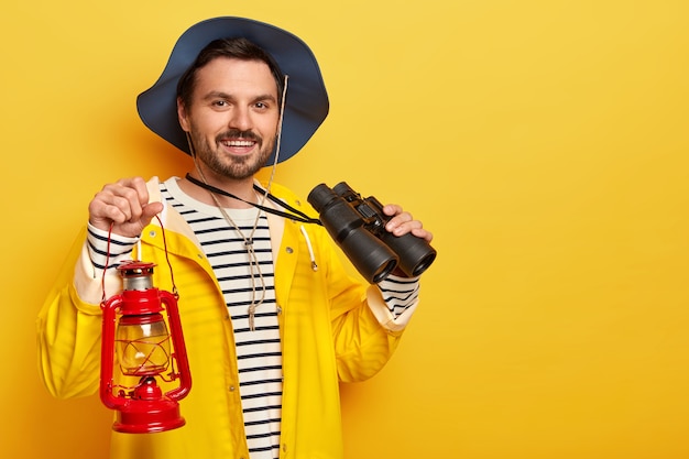 Free photo delighted man with stubble, wears headgear and yellow raincoat, carries kerosene lamp and binoculars, looks gladfully at camera, stands indoor