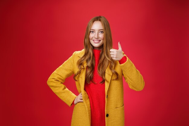 Delighted and satisfied female redhead customer showing thumbs up in like and approval pleased with great quality of new yellow coat standing over red background, smiling broadly happy.