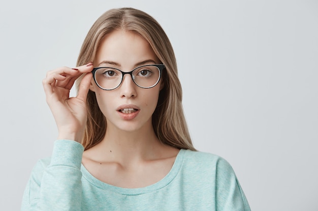 Free photo delightful pretty blonde woman in trendy eyeglasses, wears light blue sweater, stands indoors