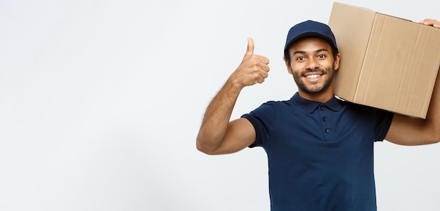 Free Photo delivery concept portrait of happy african american delivery man holding a box package and showing thumps up isolated on grey studio background copy space