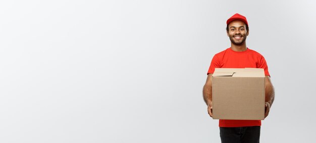 Delivery Concept Portrait of Happy African American delivery man in red cloth holding a box package Isolated on Grey studio Background Copy Space