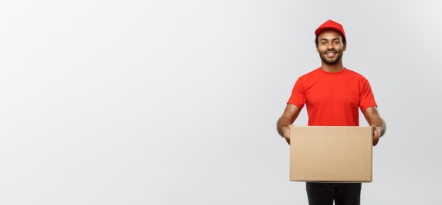 Delivery Concept Portrait of Happy African American delivery man in red cloth holding a box package Isolated on Grey studio Background Copy Space