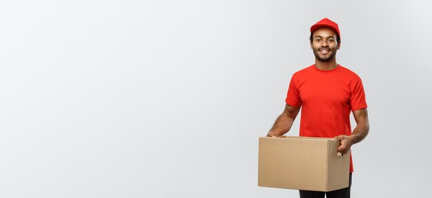 Delivery Concept Portrait of Happy African American delivery man in red cloth holding a box package Isolated on Grey studio Background Copy Space