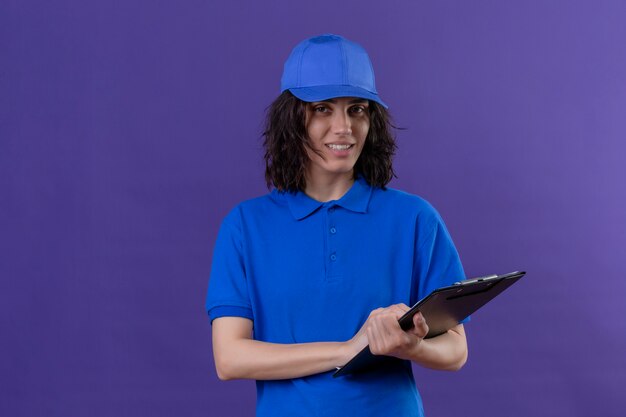 Delivery girl in blue uniform and cap holding clipboard with friendly smile standing on isolated purple