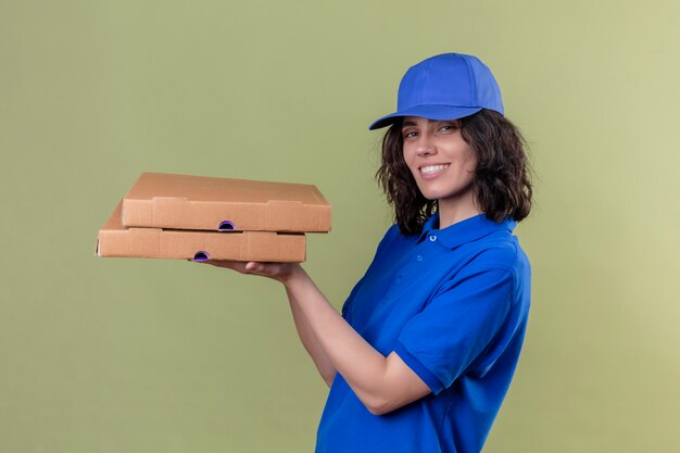 Delivery girl in blue uniform and cap holding pizza boxes looking joyful positive and happy smiling cheerfully standing over isolated green space