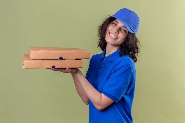 Delivery girl in blue uniform and cap holding pizza boxes looking joyful positive and happy smiling cheerfully standing on isolated green