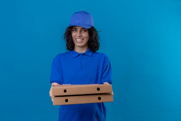 Delivery girl in blue uniform and cap holding pizza boxes positive and happy smiling friendly standing on isolated blue