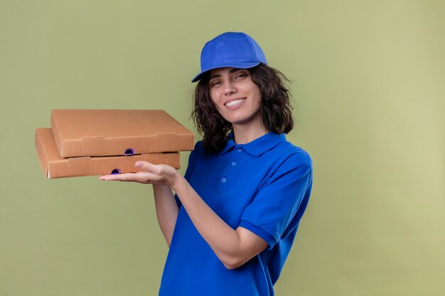 Delivery girl in blue uniform and cap holding pizza boxes smiling friendly and confident standing over isolated olive color space