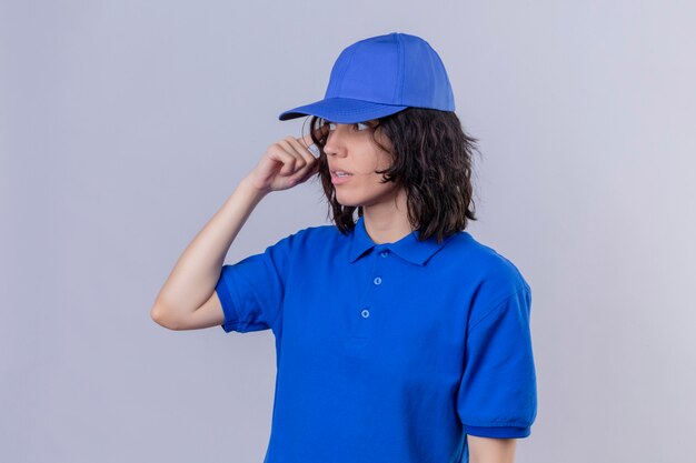 Delivery girl in blue uniform and cap looking away with confused expression standing on isolated white