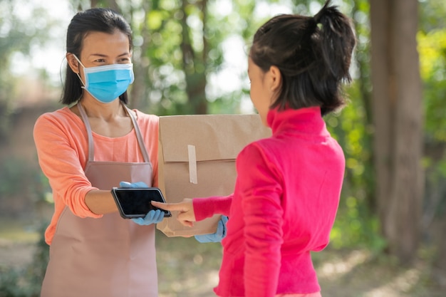 Delivery, mail, people and shipping concept.Young woman sign in digital mobile phone after receiving parcel from courier at home. from delivery man during COVID-19 outbreak.